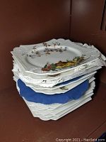 Stack of eight square porcelain plates with embossed borders and various hand-painted designs, partially showing detailed decorating and lace doilies beneath.