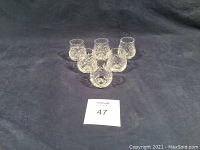 Photo showing six clear crystal shot glasses arranged in a circle on a black cloth background.