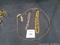 Photo showing a silver-toned large link necklace, gold-toned necklace, silver fine small link chain necklace, additional silver small chain necklace, silver-toned bracelet, gold-toned lapel pin, and small stud earrings on blue background.