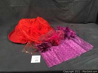 Photo showing red chiffon dress hat, red straw hat with red flower band, and purple fascinators with netting and flowers all placed on a dark fabric background.