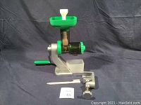 Front angled view of manual juicer showing green hopper, crank handle, clamp, and cleaning brush on a grey backdrop.