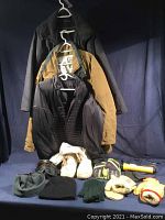 Three jackets (black Bill Blass, tan Geraldo Arctic Discovery Parka, black MEC Thermo Lite jacket) hung on hangers, winter boots, gloves, hats, and a yellow flashlight on a blue fabric background