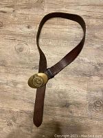 Brown leather belt laid out on wood floor, showing entire length and oval gold buckle.