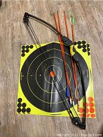 Full set including black recurve bow, four wooden arrows, and three yellow and black target sheets laid out on wood floor.