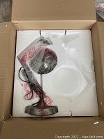 Desk lamp securely packed in white foam inside cardboard box with lamp components wrapped in plastic.