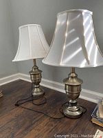 Full view of two metal table lamps with white fabric bell-shaped shades placed on wood floor in front of wall corner.
