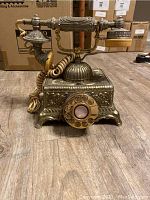 Front view of vintage telephone reproduction showing rotary dial, handset cradle, and ornate metallic body with floral detailing.