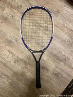 Full view of blue, white, and black tennis racket with strings and black grip on wooden floor.