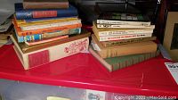Two stacks of assorted hard and soft cover books arranged on a red plastic container lid. Visible titles include One Flew Over the Cuckoo's Nest and classic novels. Books show signs of wear and age, some with illustrated covers.