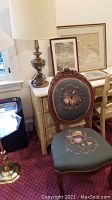 Photo of vintage floral embroidered carved wooden chair with upholstered seat and back, sitting on a red patterned carpet beside a metal lamp with a cream shade.