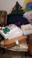 View of boxed Christmas tree, porcelain house model in styrofoam tray, glass ornaments in striped box, and other items stacked on chairs and boxes.