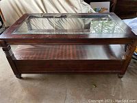 Angled view of wooden coffee table showing glass top and lower shelf design with visible scratches on wood surface and glass insert.