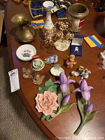 Overview of assorted figurines and floral decor on table including brass ware, a small spouted vase, trinket plate, porcelain flowers, and multiple small figurines including Hummel style children.