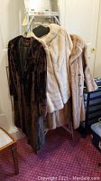 Three vintage fur coats on hangers. One dark brown beaver coat with visible damage, two light-colored mink coats. Coats vary in length from waist to mid-thigh. Photographed indoors on wooden easel hanger.