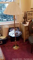 Full view of metal floor lamp with attached glass top table, white pleated lampshade, brass finish, set next to a window and furniture.