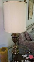 Polished metal lamp with urn-style base and white drum shade on a glass side table with candle and sofa visible.