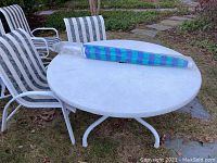 Photo of round white patio table with folded umbrella on top, set outdoors on grass, surrounded by two striped chairs
