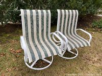 Pair of outdoor patio high back swivel chairs with white metal frames and green and white vertical striped fabric seats and backs, shown outdoors on grass with trees in background.