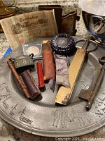 Full view of pewter tray with all items arranged on it including barbers tool, skeleton key, coin, ribbon, scissors, brush, bell, book, lipstick case and German banknote