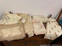 Overview of folded vintage linens including tablecloths, placemats, and napkins in neutral shades with embroidery and lace details on top of wooden furniture.