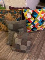 Photo shows 6 throw pillows in various colors and patterns, placed on floor against wall. Patchwork pillows, colorful geometric pillow, floral sun gold design pillow, and solid purple pillow visible.