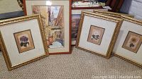 Six pieces of wall art arranged on carpet floor, showing four floral prints in ornate gold-tone wood frames and two other framed prints including a street scene and a nature theme.