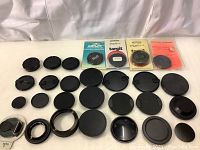 A pile of about 30 various black camera lens caps, rear caps, and lens adapters, with three complete Targit brand packages in original packaging at the top