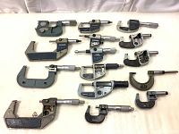 Photo showing 17 micrometers of various types and sizes arranged on a white surface, including digital, mechanical, and dial gauge models with visible signs of wear, missing parts, and some rust.