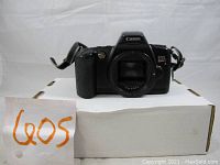 Front view of Canon EOS Rebel X film camera body with no lens, mounted strap, sitting on white box with orange writing 'EOS'.