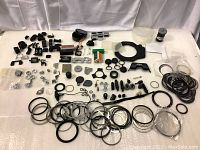 Wide view showing numerous small camera parts including rings, caps, batteries, and other mechanical components laid out on a surface.