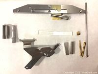Full overview of all parts including long metal tool, cylindrical handle, small brass parts, acrylic piece, rectangular metal blocks, and angled assembly component.