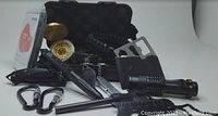 The photo shows a collection of survival and emergency tools arranged in front of an open black carrying case with foam padding. Items include a brass compass, black tactical flashlight, multi-tool credit card, emergency whistle, fire starter, window break tool, two carabiners, and a small tactical pen or knife.