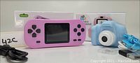 Photo showing a pink handheld game console with directional and action buttons, a blue kids video camera with a blue wrist strap, their respective boxes, and cables, all new in box.