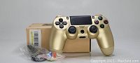 Metallic gold PlayStation 4 DualShock 4 controller displayed with original cardboard box and included accessories such as USB charging cable and thumb grips.