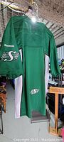 Back view of green Saskatchewan Roughriders jersey by Reebok, hanging indoors with light overhead.