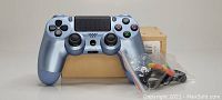 Titanium blue PS4 wireless controller shown resting on its original box with plastic bag containing various cables and accessories in front.