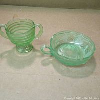 Photo showing two green Depression glass bowls, one with two handles on a pedestal and one with one handle and scalloped edge with grapevine embossing.