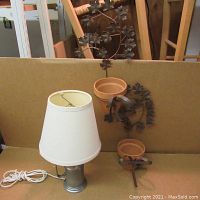 Lamp with white fabric shade and brushed metal base alongside two metal hanging plant holders with terracotta pots and leaf designs on a brown background.