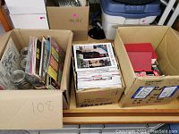 Three cardboard boxes: one filled with vinyl LPs, another with magazines, and a third with books and boxed items.