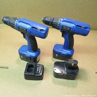Photo showing two blue cordless drills with battery packs and a black charger base.