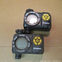 Front view of two black Melnor sprinkler control units showing circular dial with instructions and yellow buttons on right side