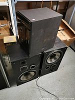 Photo showing two large vintage speakers with visible large drivers and circular ports alongside a smaller unit placed on top, all exhibiting signs of use and dust.