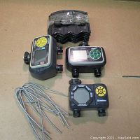 Three sprinkler control units and a set of black plastic lawn edging stakes arranged on a cardboard surface.