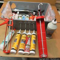 Overview photo showing multiple caulk tubes with various colored labels and two manual red caulking guns on work surface, clear plastic bin in background