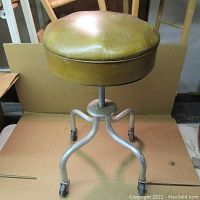 Front view of vintage adjustable rolling stool showing greenish-yellow vinyl padded seat, metal four-legged base with casters.