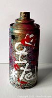 Front view of painted spray can showing detailed layered paint splatters and white/red graffiti markings.