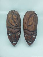 Front view of two carved wooden West African style masks with detailed facial features and hair carved in relief, eye and mouth holes painted white
