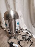 Closed front view of one stainless steel commercial tea brewer with attached power cord and European plug.