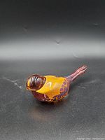 Side angle of the glass bird figurine showing the rounded amber-yellow body with brown and red spotted details and the elongated red tail