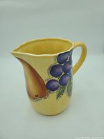 Yellow ceramic jug decorated with painted fruit including grapes and a pear, scalloped rim and handle visible.